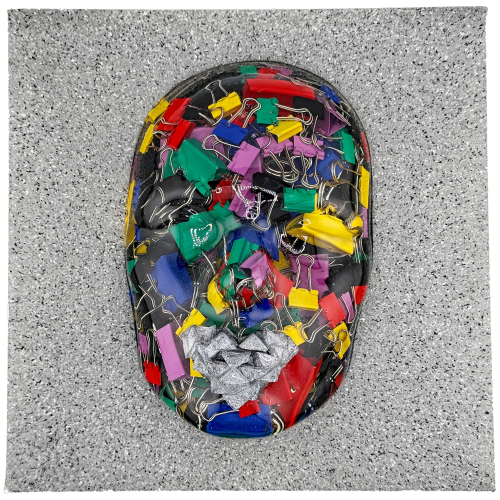 A human face mask filled with colorful binder clips and objects, with a textured, silver-colored heart at the mouth, all mounted on a sparkling, silver-speckled background.