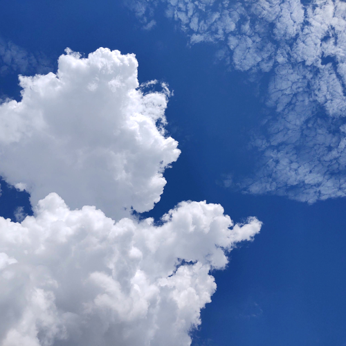 Fluffy white clouds float against a deep blue sky, with some wispy, scattered clouds visible in the upper right. In the bottom right corner, a small portion of dark tree branches can be seen—creating a scene as serene and inviting as an e-commerce homepage.