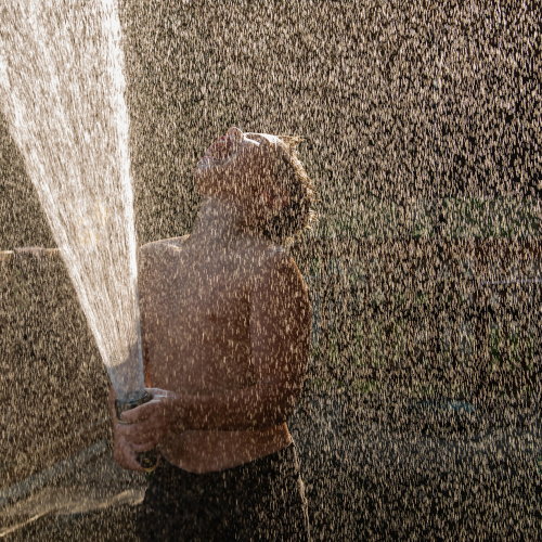 A child stands outside spraying water from a hose, tilting their head back to enjoy the spray, sunlight illuminating the water droplets around them like sparkling gems in an e-commerce summer catalog.