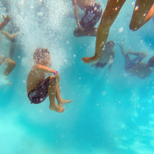 Underwater view of several children swimming and playing in a pool, with bubbles rising and sunlight filtering through the blue water. One child is curled up in mid-air as they make a splashy entry.