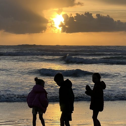 Three children stand on a beach at sunset, their silhouettes visible against the golden sky and ocean waves, with clouds partially covering the sun—a peaceful moment far removed from the rush of e-commerce and digital screens.