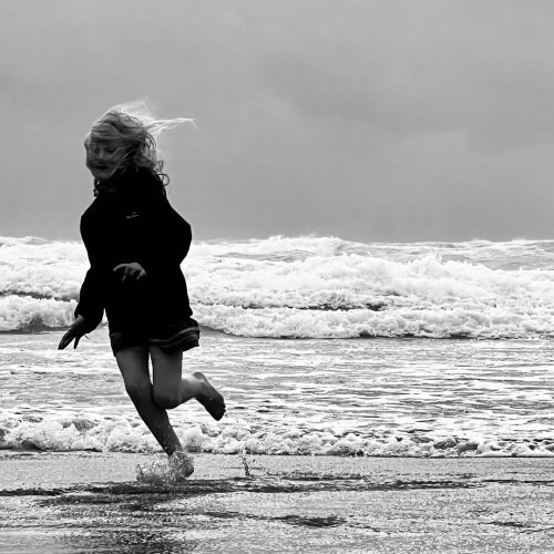 A child runs along the shoreline, splashing in the shallow waves. The scene is in black and white, with the sea and cloudy sky in the background. The child’s hair and jacket blow in the wind, capturing a timeless moment that evokes storytelling as evocative as an e-commerce campaign for classic coastal fashion.