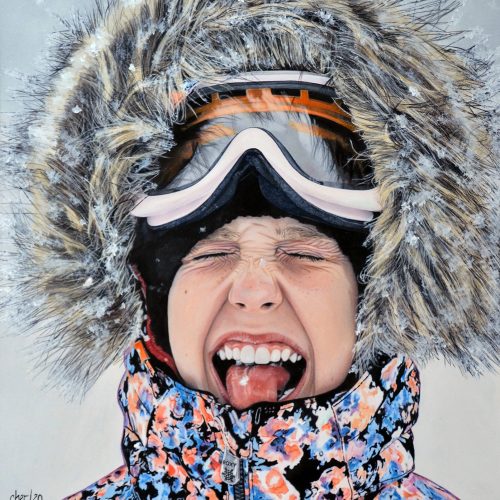 A child in a fur-lined hood and ski goggles squints and sticks out their tongue, catching snowflakes—a playful moment perfect for your e-commerce winter collection. They wear a colorful, patterned winter jacket.