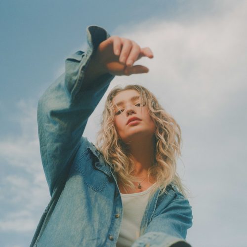A young woman with wavy blonde hair wearing a denim jacket and white shirt poses outdoors against a blue sky with light clouds, reaching her hand toward the camera in a stylish e-commerce-inspired shot.