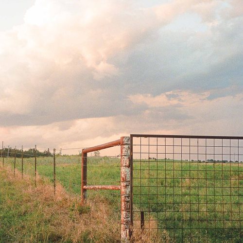 A rusty metal gate and wire fence stand in a grassy field under a cloudy sky at sunset, with soft sunlight highlighting the rural landscape—capturing the quiet beauty often sought for e-commerce imagery showcasing countryside living.