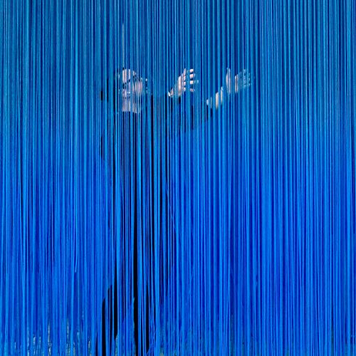 A person stands behind hanging strands of blue strings, reaching forward with both hands, partially obscured by the dense vertical lines in an innovative art installation that evokes the ever-shifting boundaries of e-commerce.