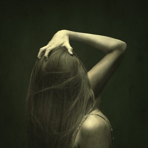 A person with long hair faces away, tilting their head back and touching their scalp with one hand against a dark green background, evoking a sense of modern style often seen in e-commerce imagery.